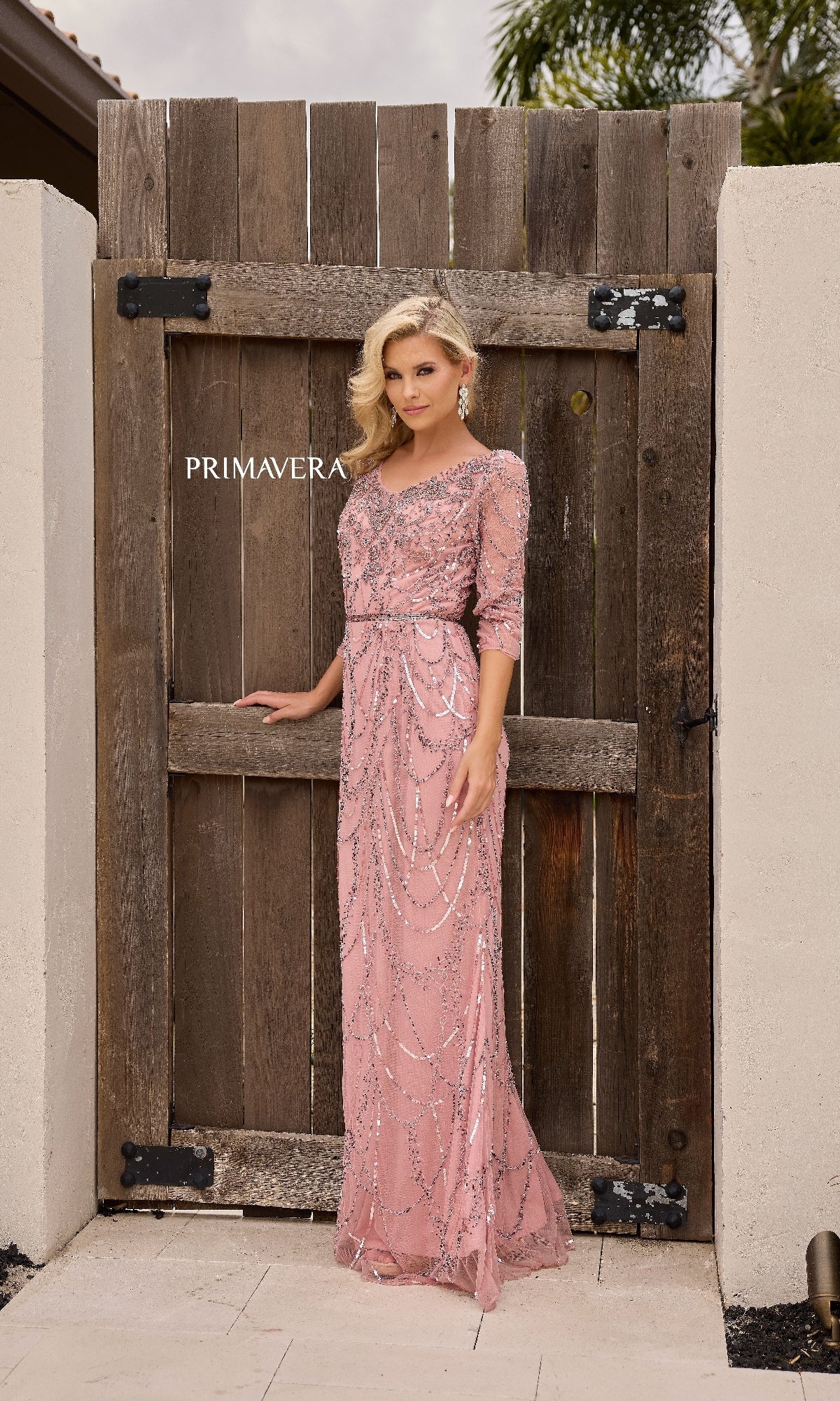 A woman stands by a wooden gate in the Primavera 13156 long formal dress, featuring a light pink hue, beaded waistline, and three-quarter sleeves.